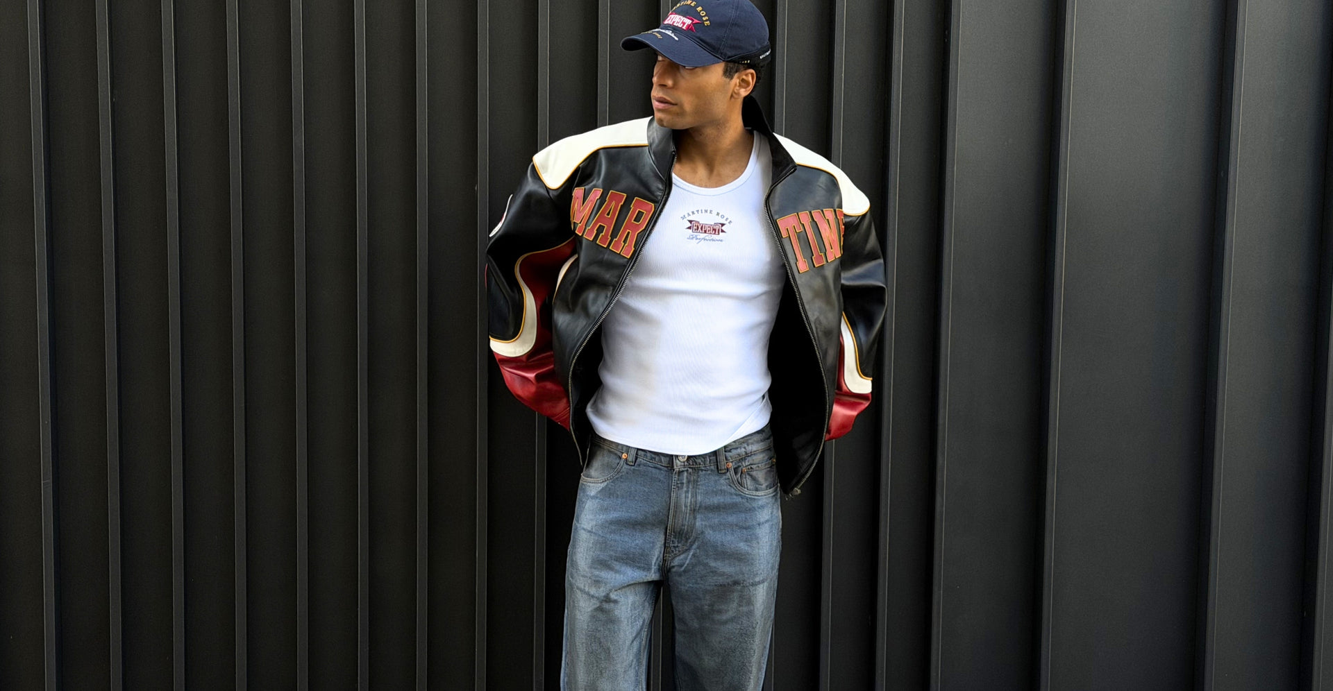 Bold men's outfit featuring a black, white and red Martine Rose leather jacket with logo, a white Martine Rose logo top, jeans and a blue cap.