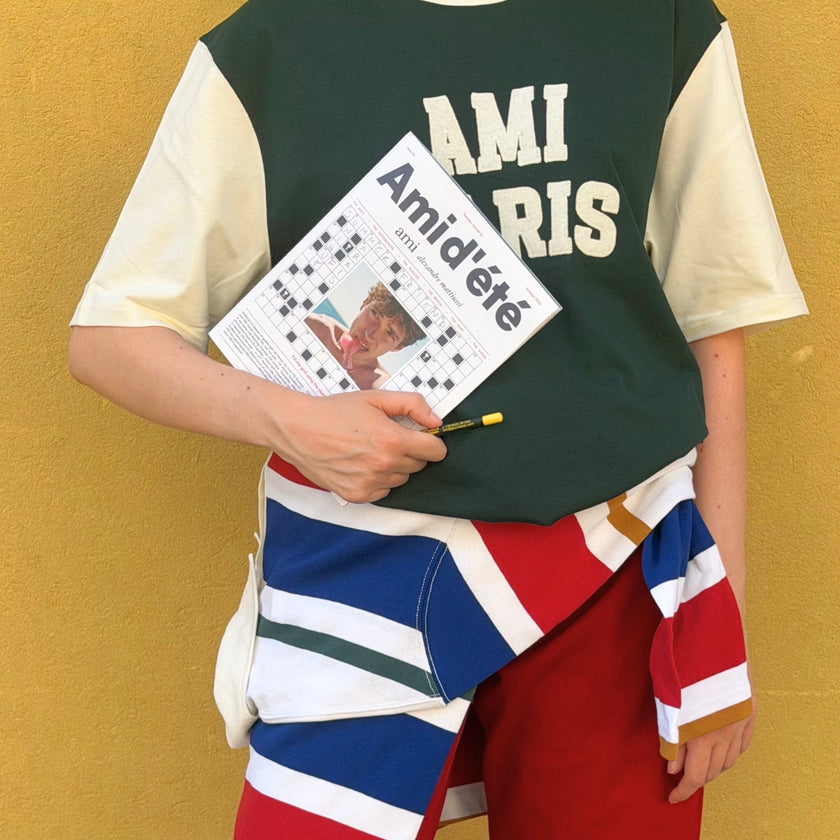 Men's Fall Winter 2026 new arrivals featuring an AMI Paris look with red jeans, a forest green logo T-shirt and a multicolor striped polo tied around the waist.