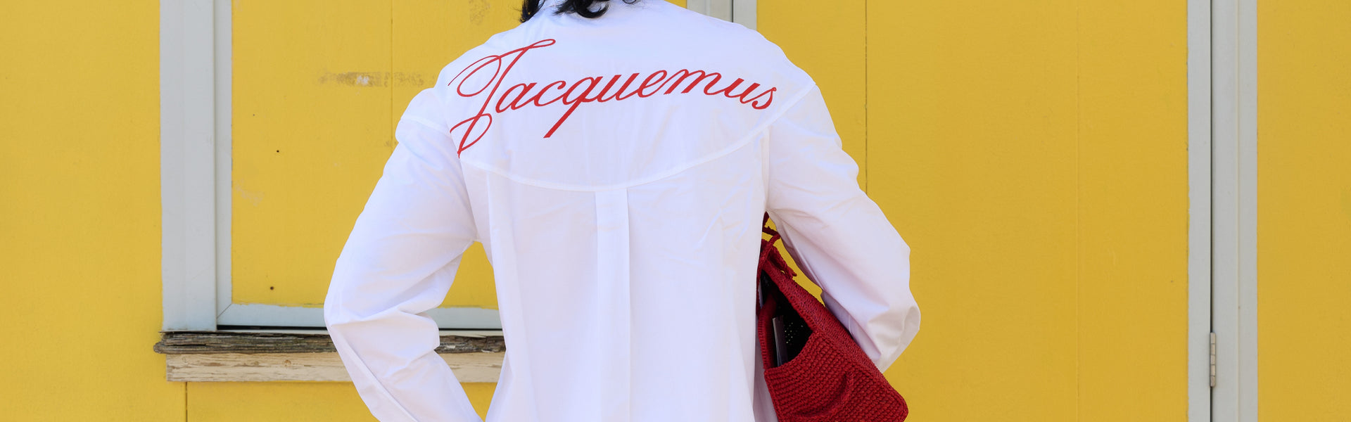 Back view of man wearing white Jacquemus shirt with red logo and holding red bag